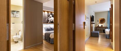 A hotel room with a wooden floor, a bed with a gray headboard, a bedside table with a lamp, a small desk with a chair, and a bathroom with a sink and toilet.