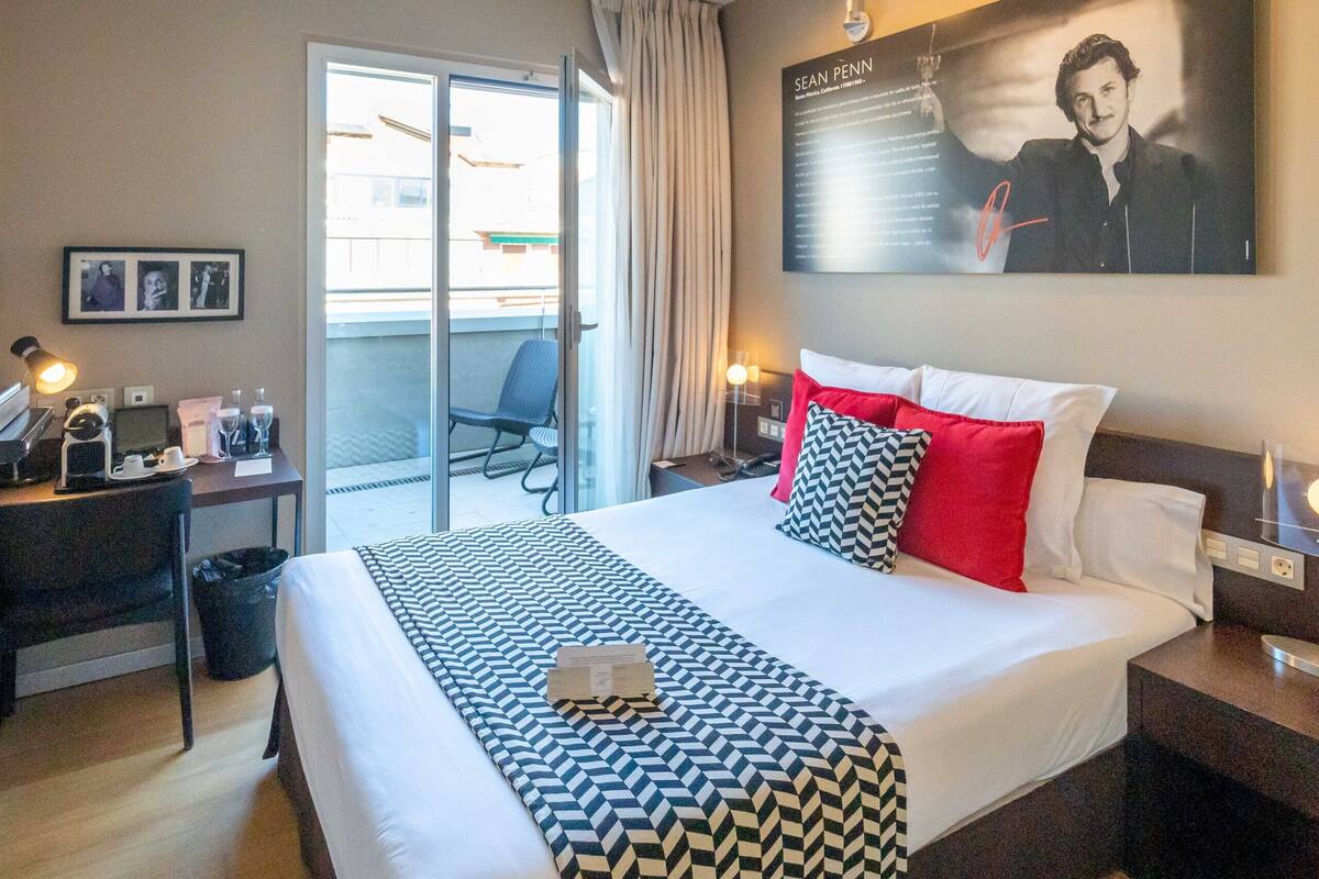 A hotel room with a large bed, a desk with a lamp, a chair, and a sliding glass door to the balcony.