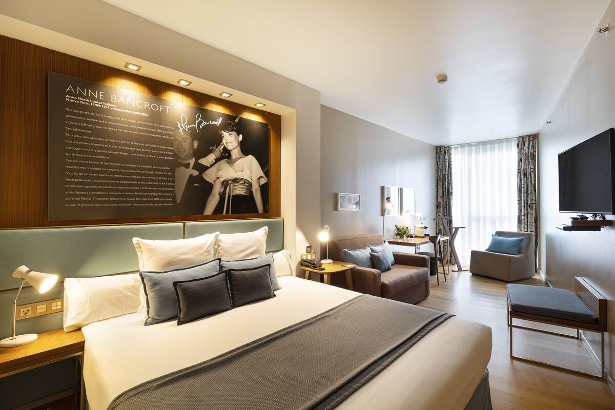 A modern hotel room with a large bed, a desk, a chair, a TV, and a wall with a poster.