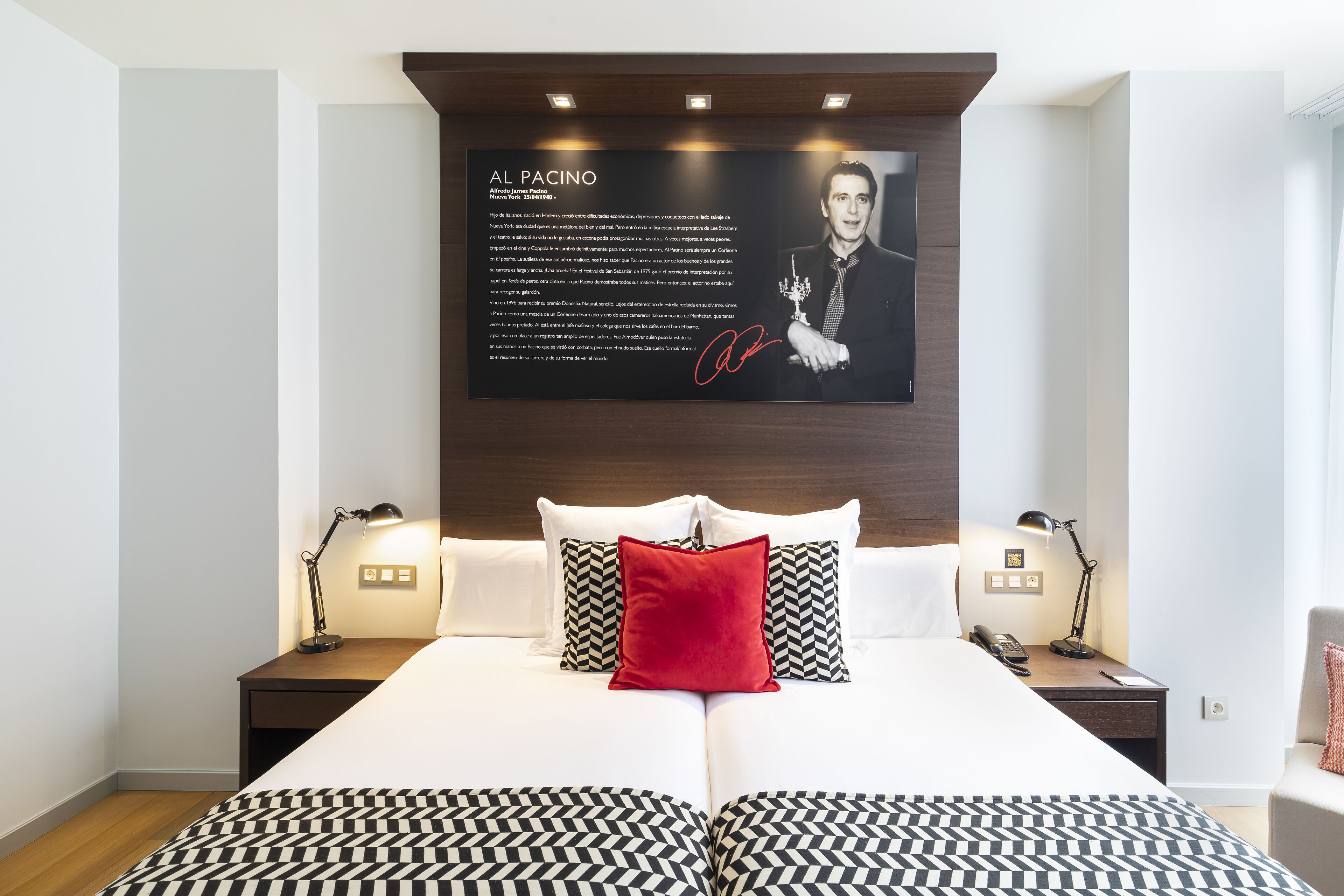 A hotel room with a large bed, two bedside tables, a nightstand, a red pillow, and a framed picture on the wall.