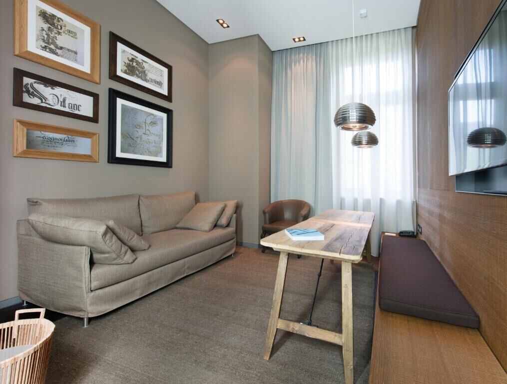 A modern living room with a sofa, a table, and a chair. There are framed pictures on the wall and a window with curtains.