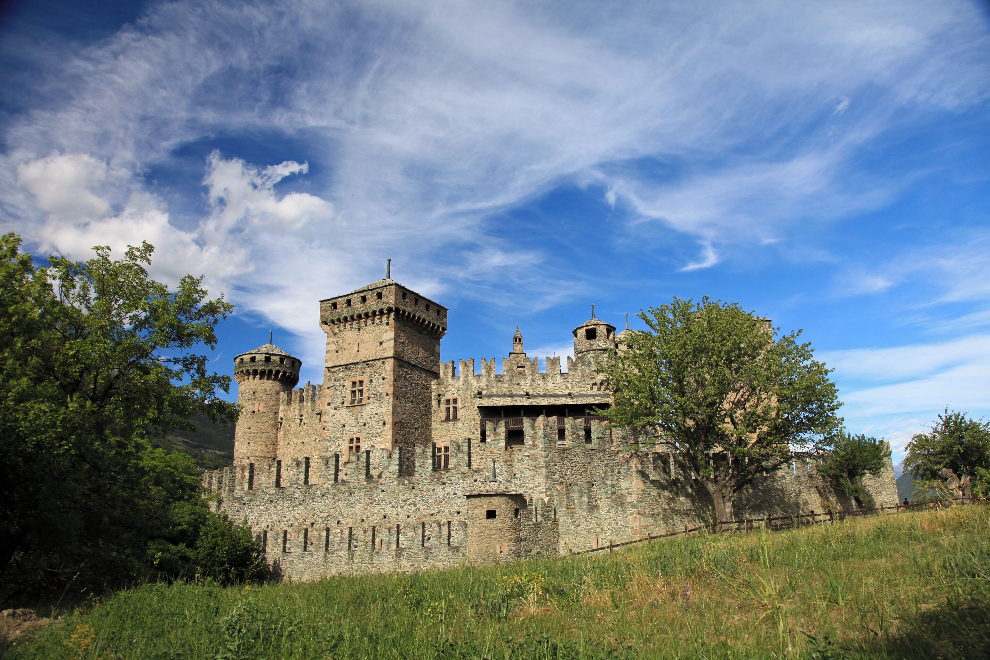 A historic stone castle with multiple towers and battlements, surrounded by lush greenery.