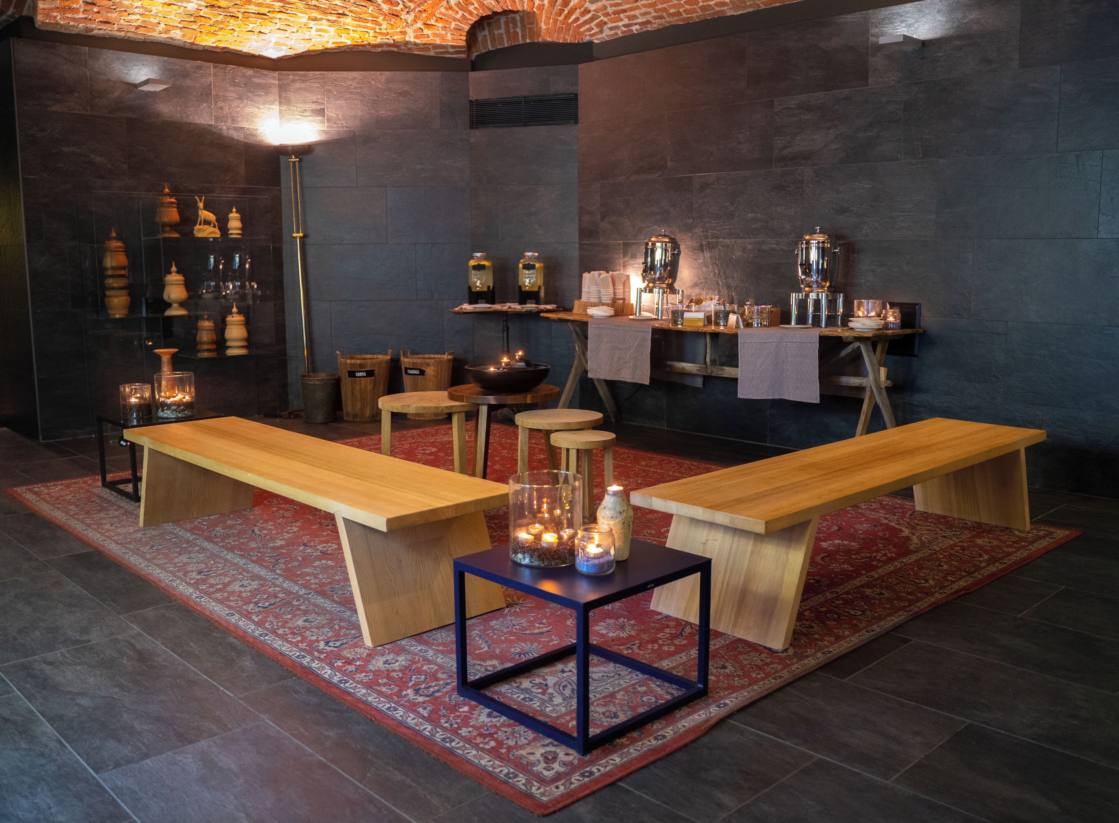 A modern interior with wooden benches, a central table, and a bar area with stools and a shelf of bottles.