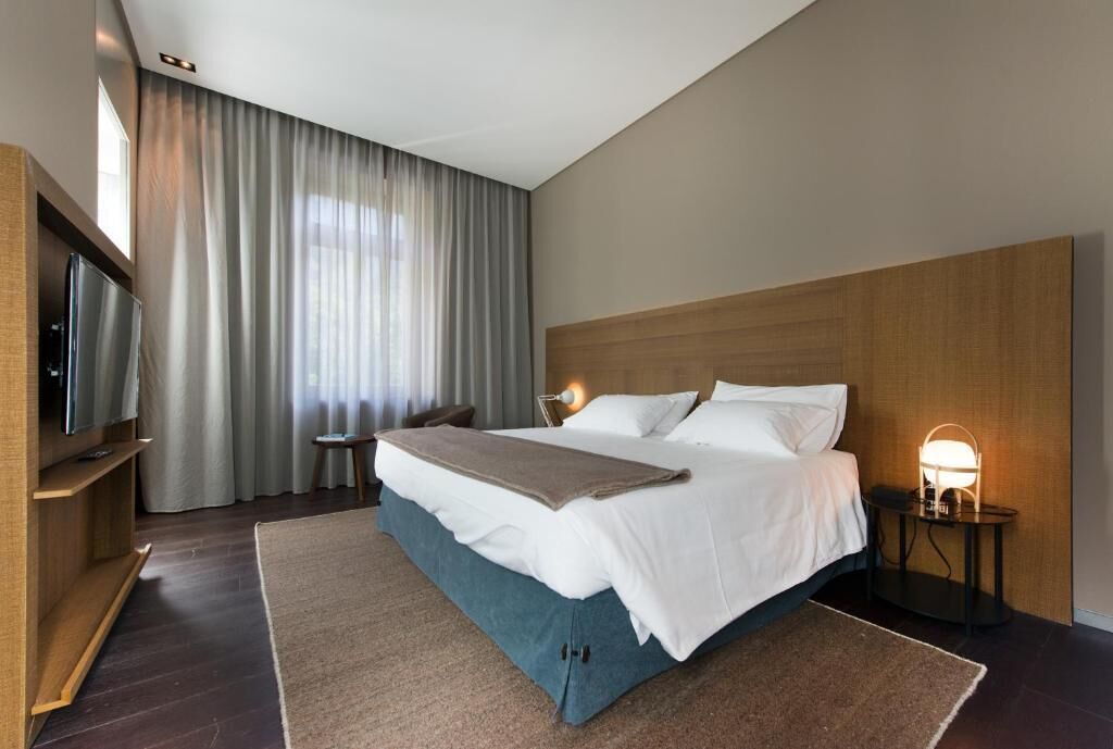 A hotel room with a large bed, a TV, a bedside table with a lamp, and a window with curtains.