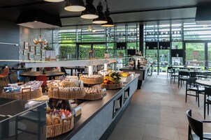Un café moderno con una barra larga que exhibe diferentes alimentos y cestas, grandes ventanales con vistas a zonas verdes, y una decoración interior limpia y contemporánea.