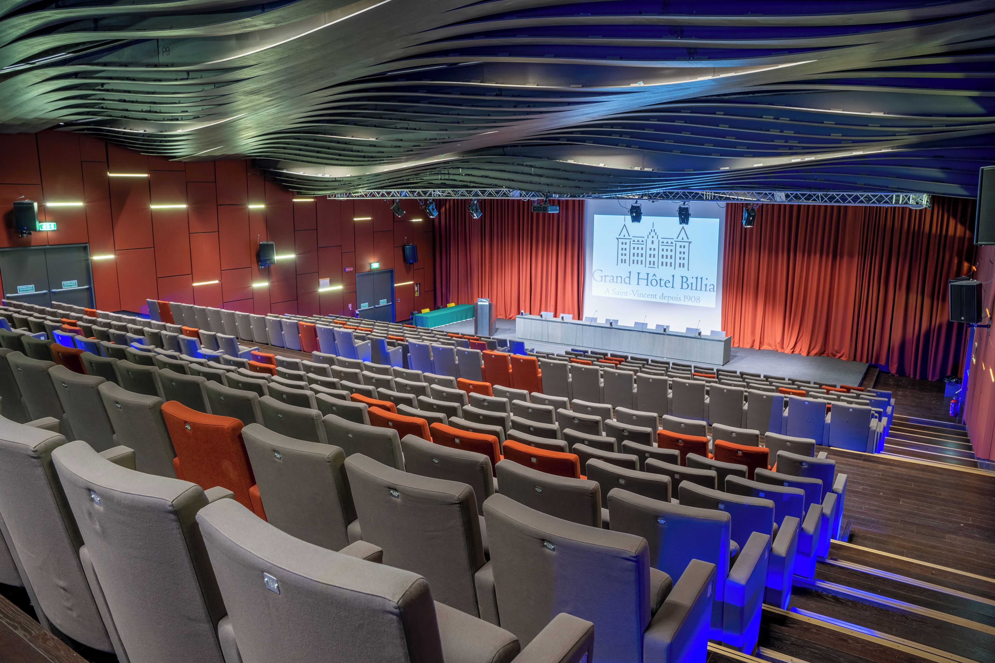 Un auditorio con hileras de asientos, un escenario con una pantalla de presentación y un telón rojo de fondo.
