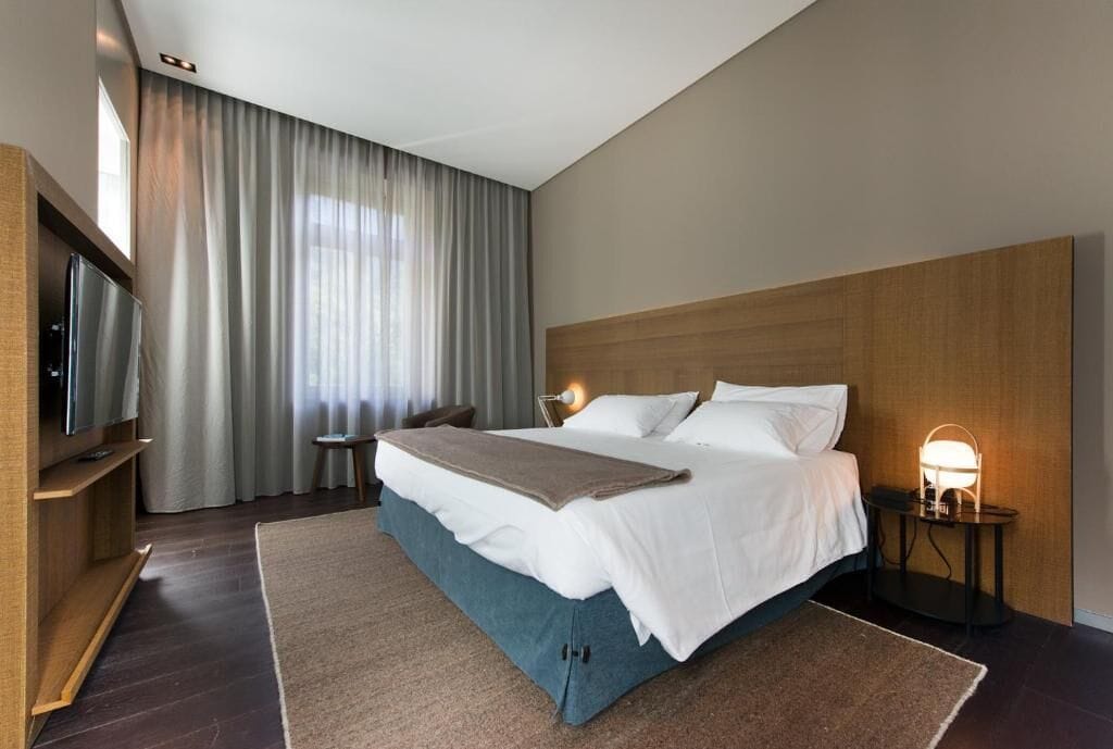 Habitación de hotel moderna con una cama grande, televisión de pantalla plana, mesita de noche con lámpara y vistas al exterior a través de cortinas translúcidas.