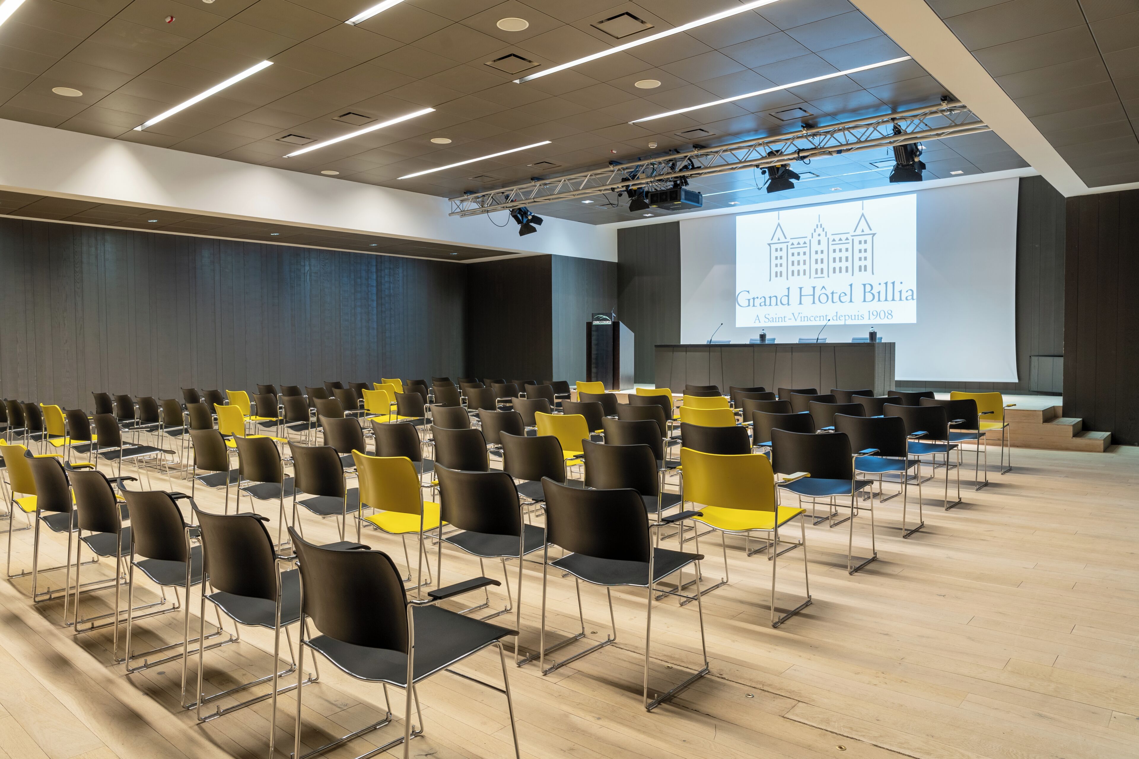 Ein Konferenzraum mit Reihen von Stühlen, einer Bühne mit einer Präsentationsleinwand und einem Rednerpult.