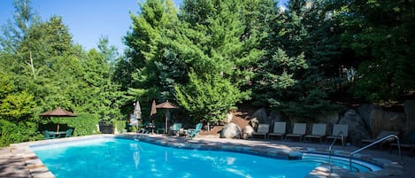 Une piscine d’un bleu éclatant, entourée de chaises longues et de parasols, avec des arbres et un mur de rochers en arrière-plan.