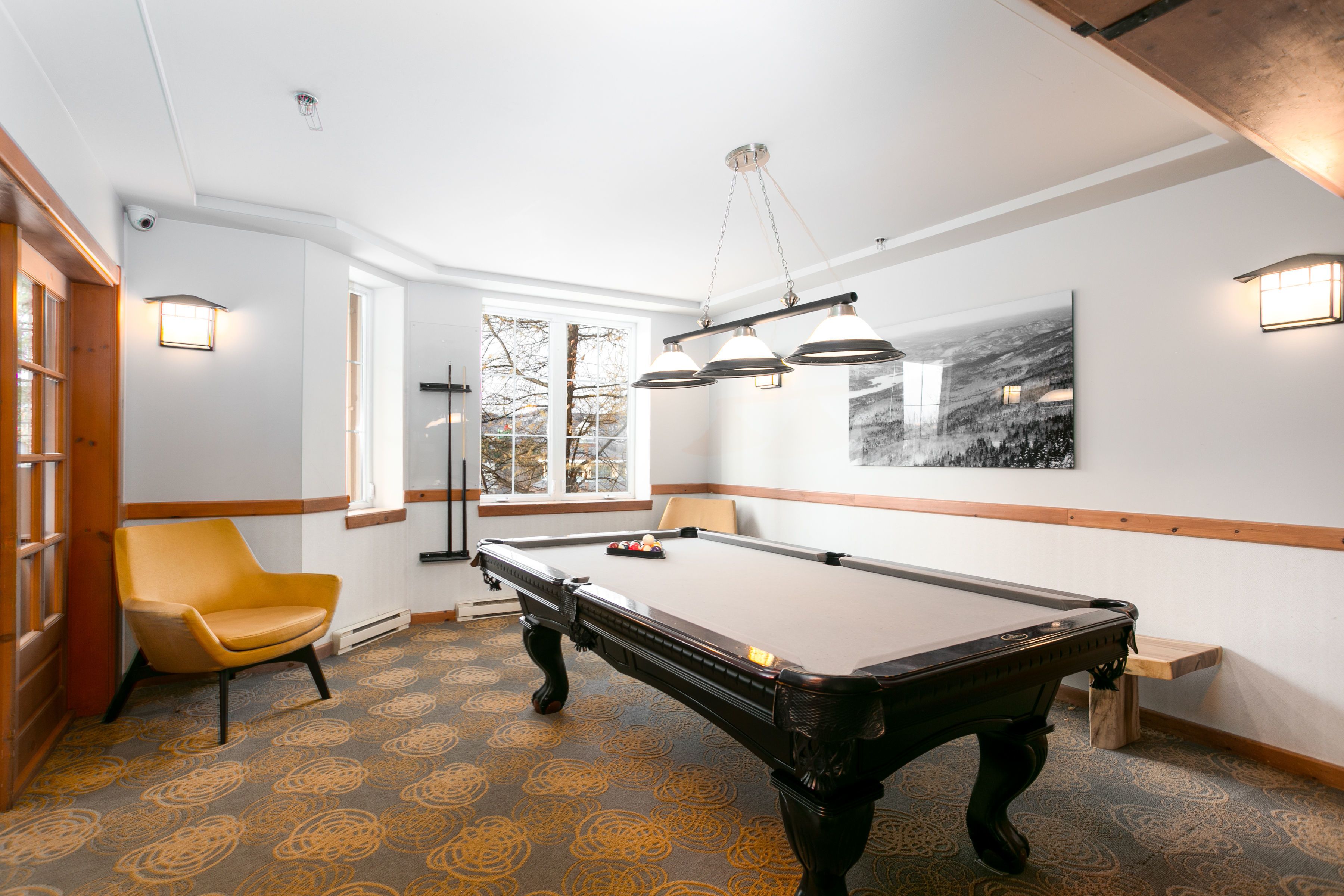 A pool table in a room with patterned carpet, a yellow chair, and a painting on the wall.