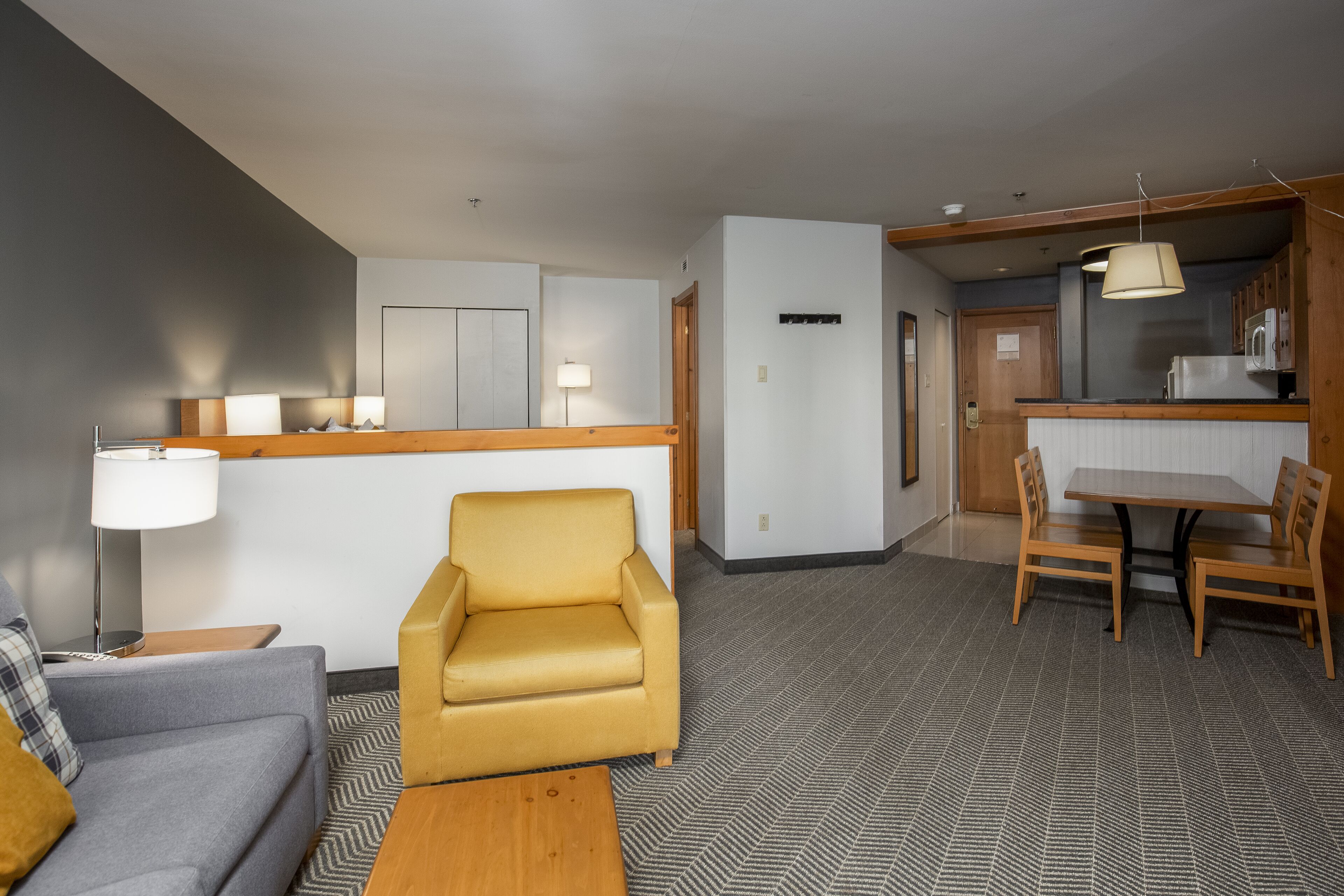 A modern hotel room with a sofa, a yellow armchair, a wooden coffee table, a dining table with chairs, a kitchen area with a microwave, and a lamp.