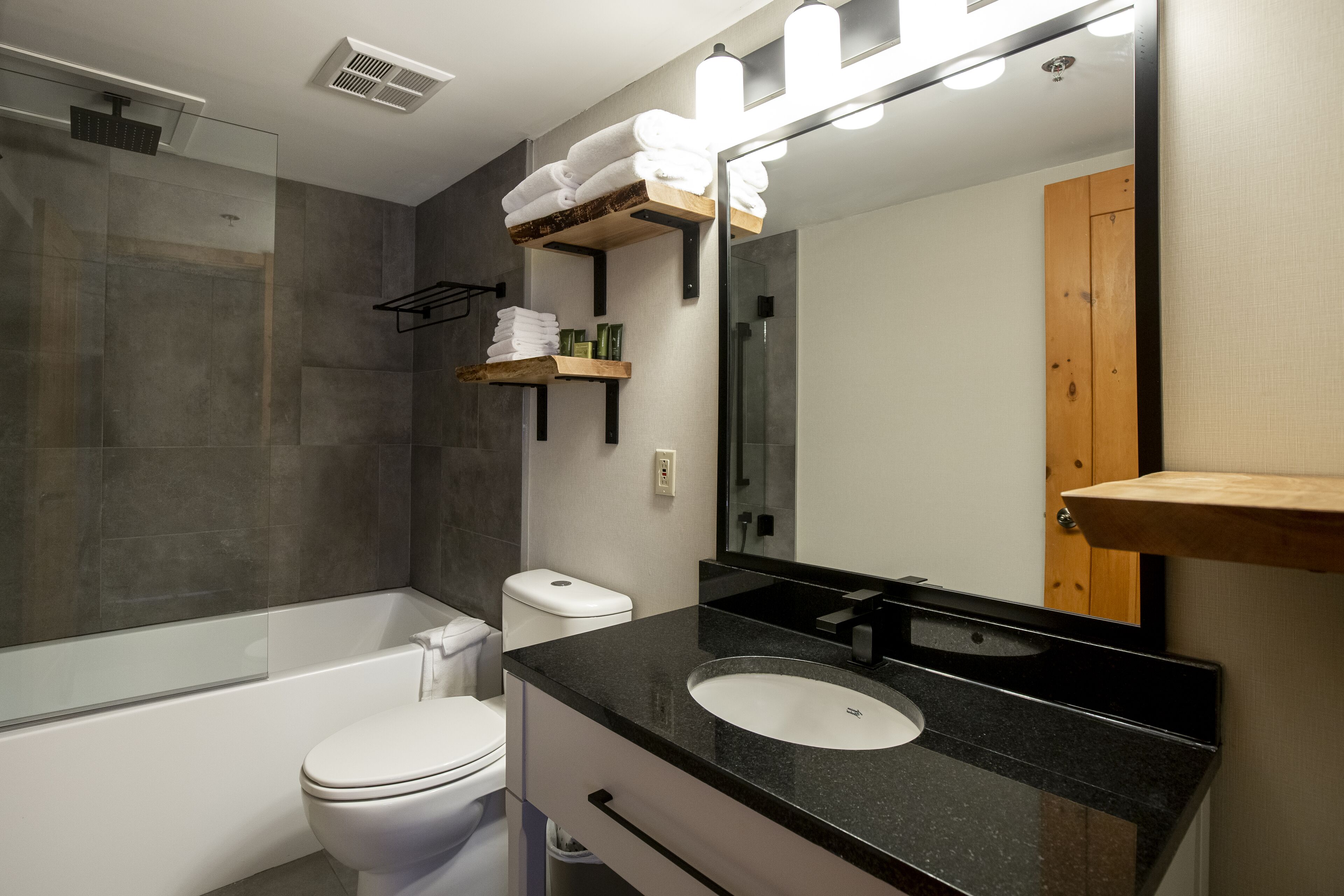 A modern bathroom with a glass-enclosed shower, a white toilet, a black sink, and a mirror with built-in lights.