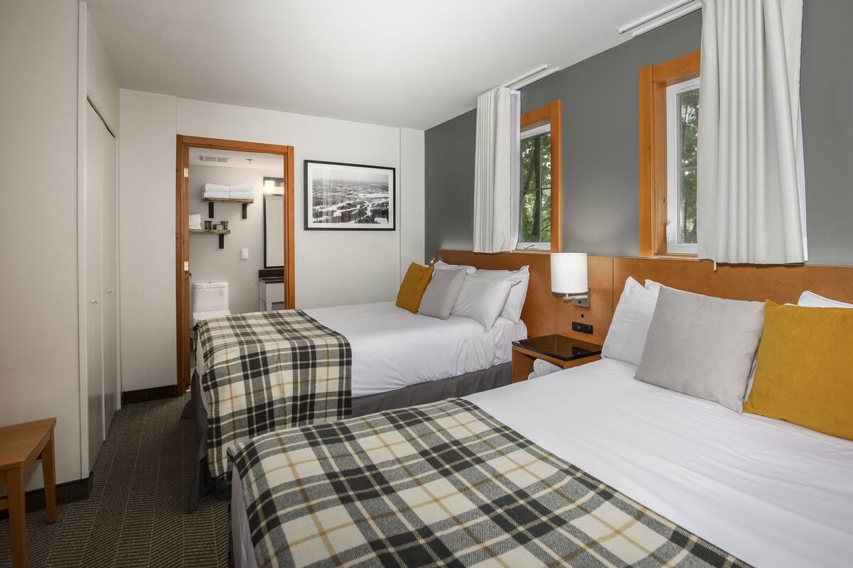 Ein Hotelzimmer mit zwei Betten, einem kleinen Tisch, einem Nachttisch, einem Fenster mit Vorhängen, einem gerahmten Bild an der Wand und einem durchgehenden Türspalt sichtbaren Badezimmer.