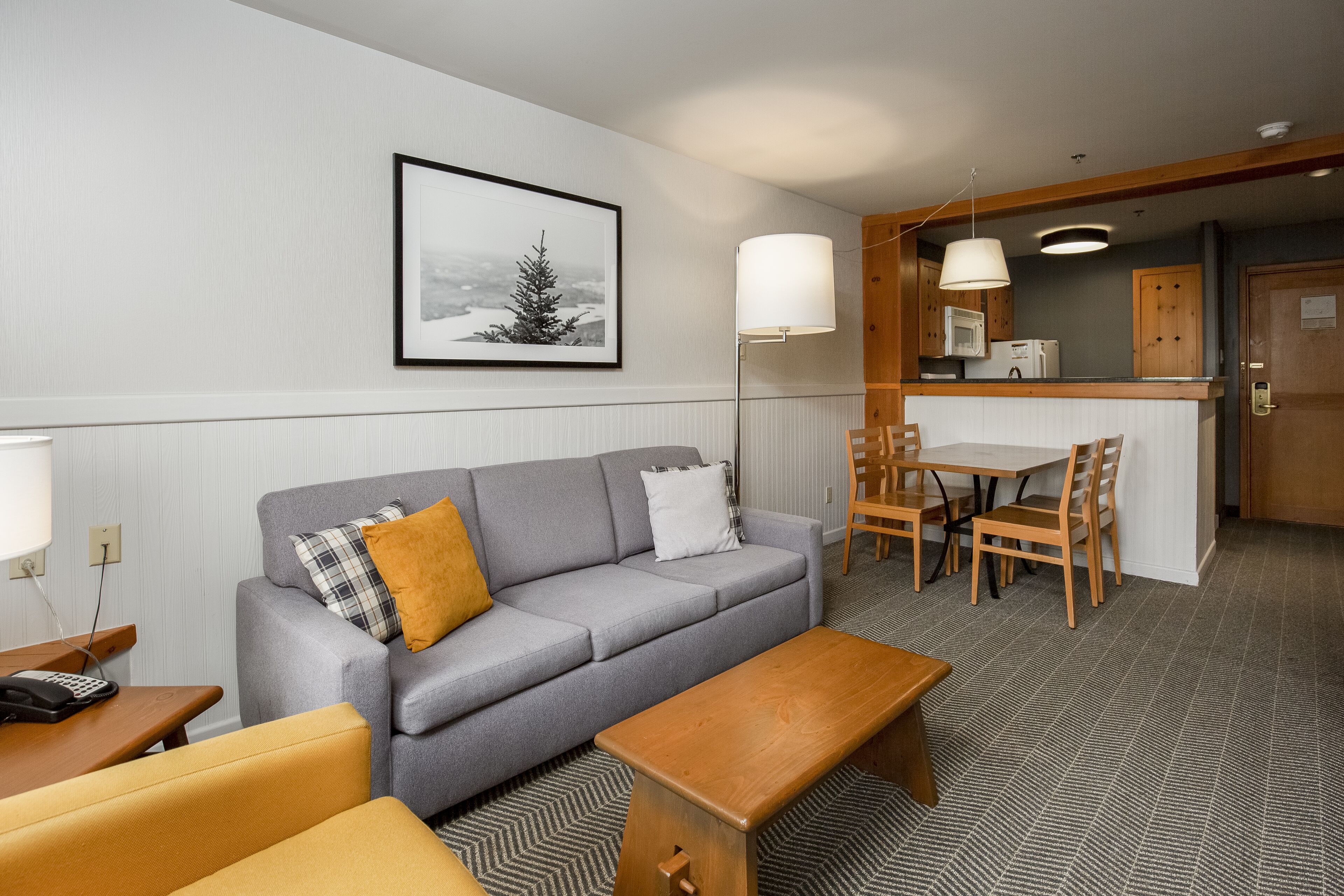 A modern living room with a grey sofa, wooden coffee table, and a dining area with a table and chairs. A kitchen area is visible in the background.