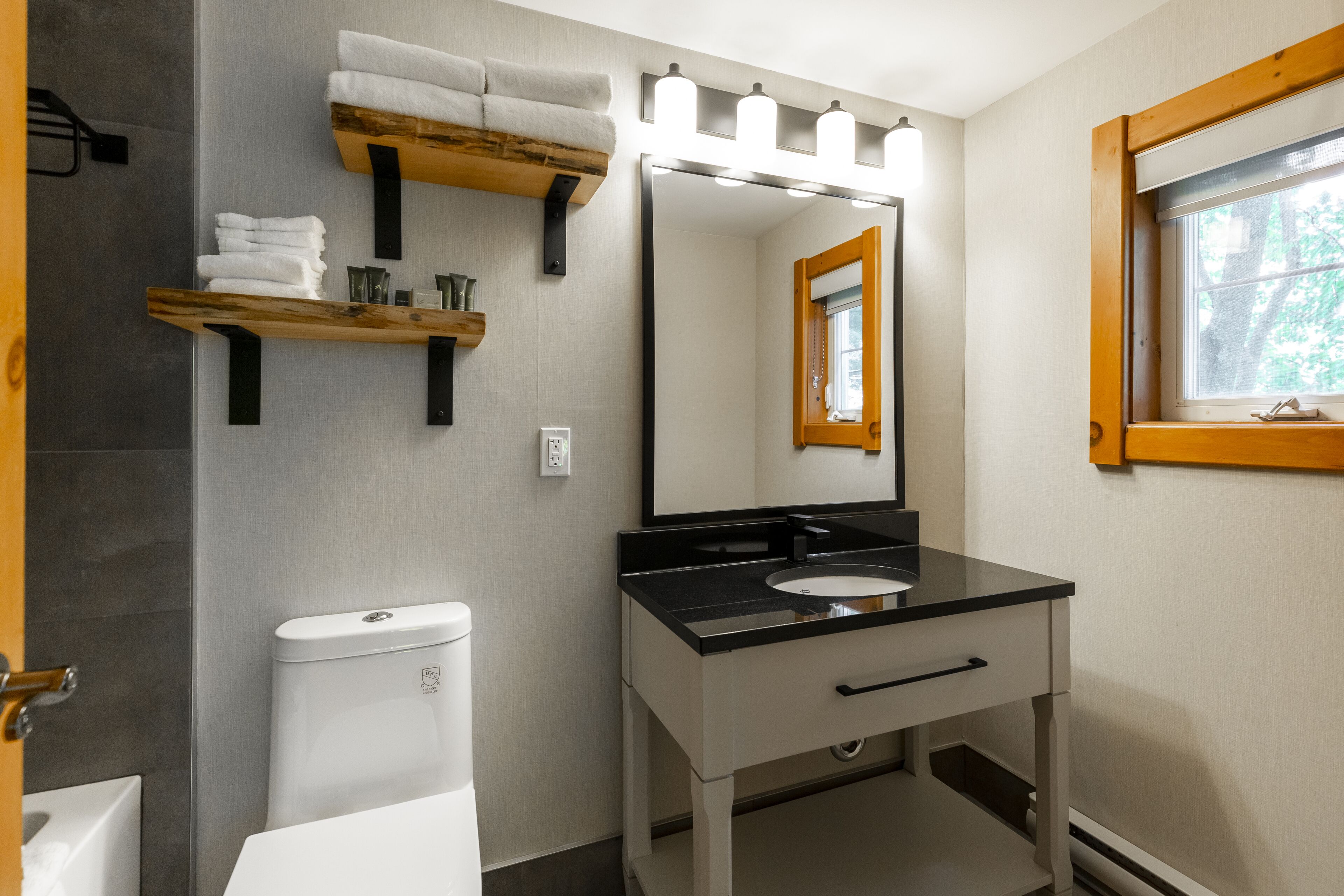A modern bathroom with a toilet, sink, and vanity. There are wooden shelves with towels and toiletries. A large mirror with lights above it is mounted on the wall.
