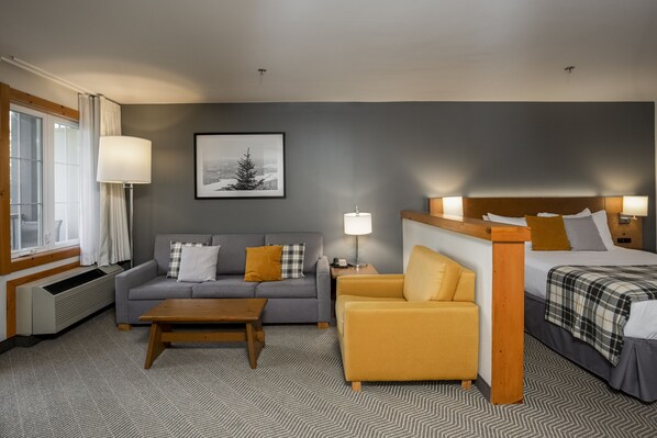 Un cuarto de hotel con un sofá gris, un sillón amarillo, cama con cabecera de madera, un cuadro enmarcado en la pared y una ventana con cortinas.