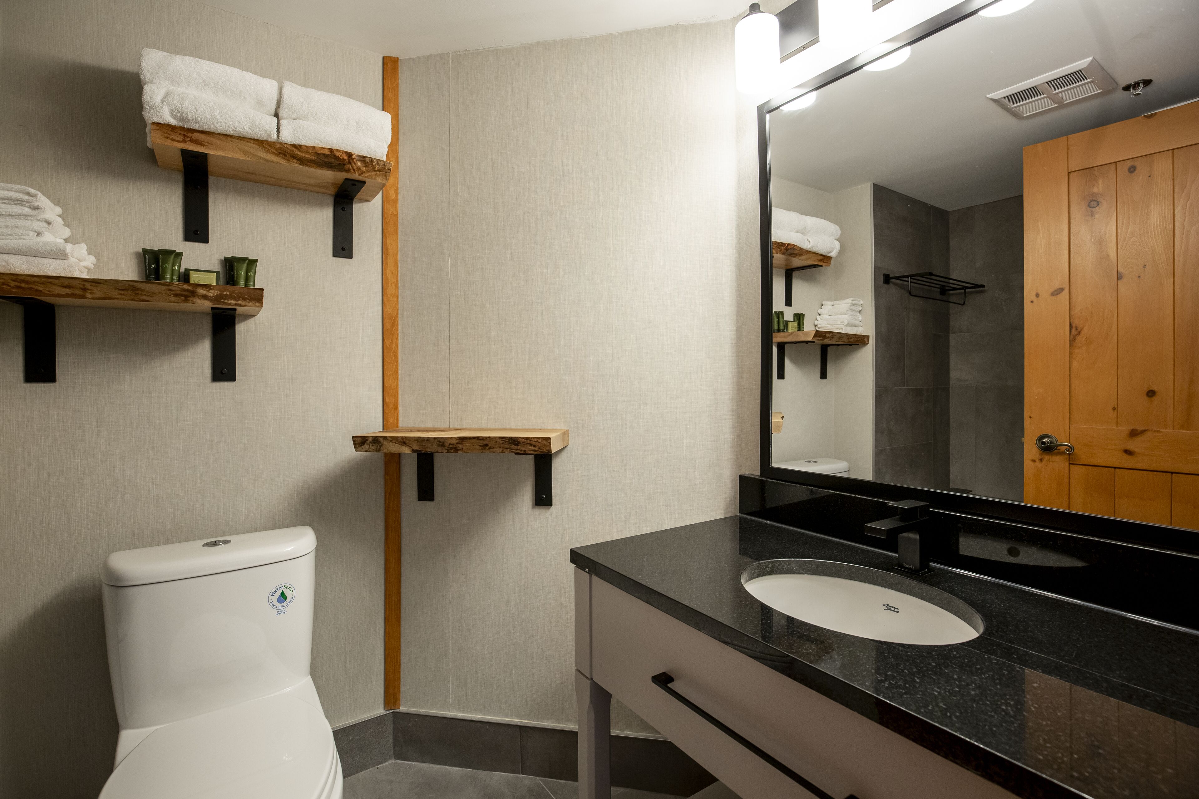 Une salle de bain moderne avec des toilettes, un lavabo et un miroir. On y trouve des étagères en bois où sont rangées des serviettes et des articles de toilette.