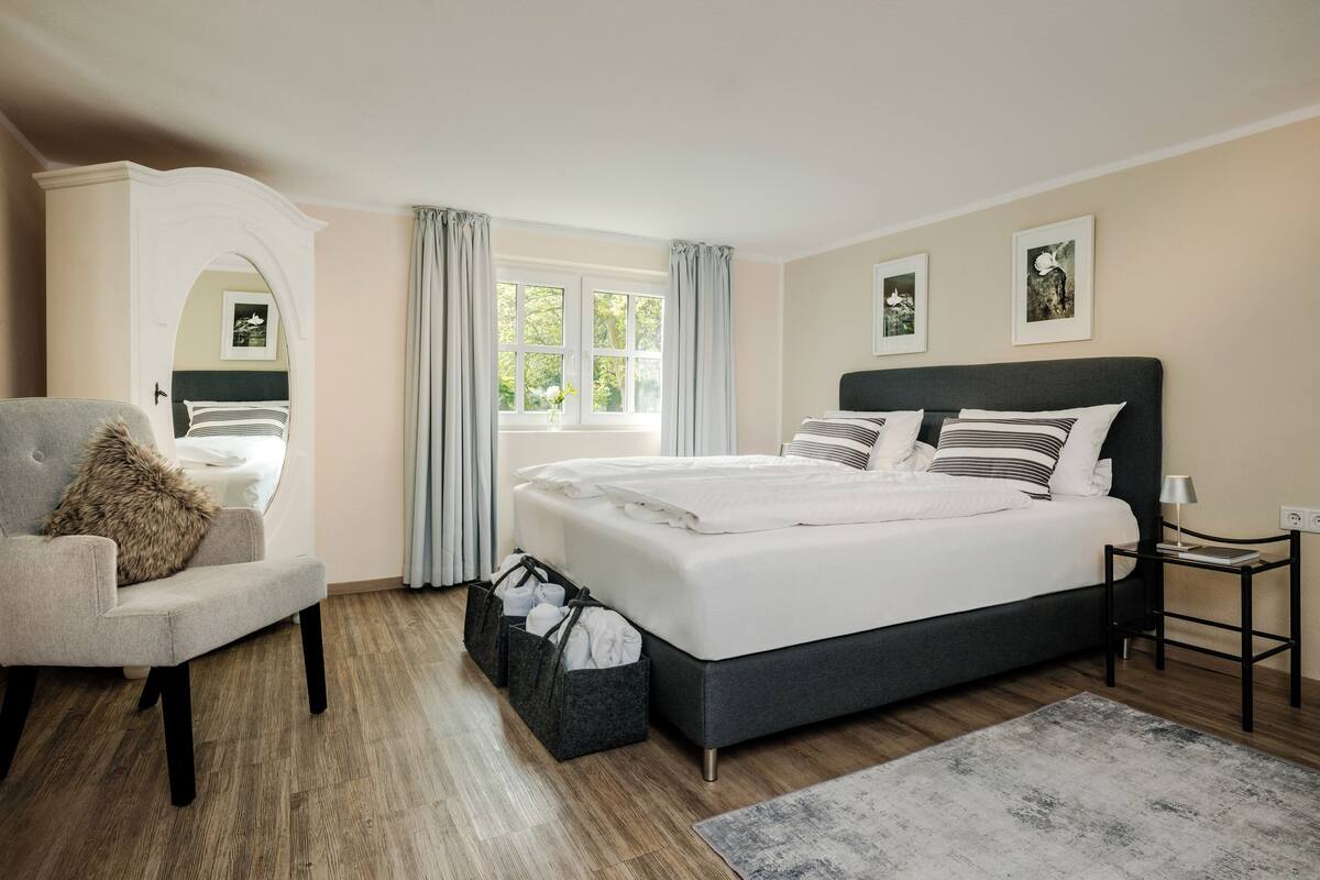 A modern bedroom with a large bed, a grey armchair, a small table, and a window with curtains.