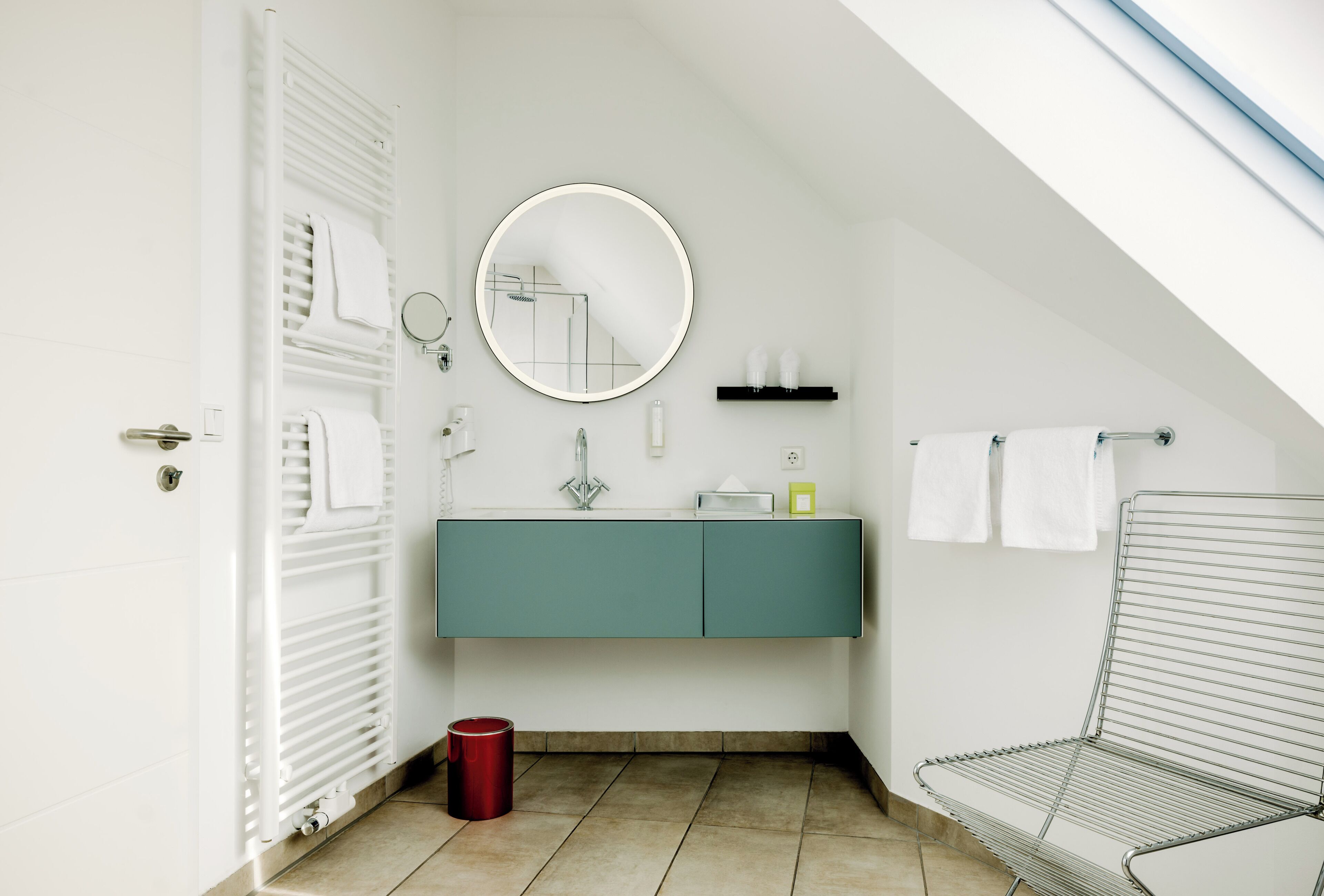 Un baño moderno con un radiador blanco, un tocador verde azulado, un espejo redondo y un cubo de basura rojo.