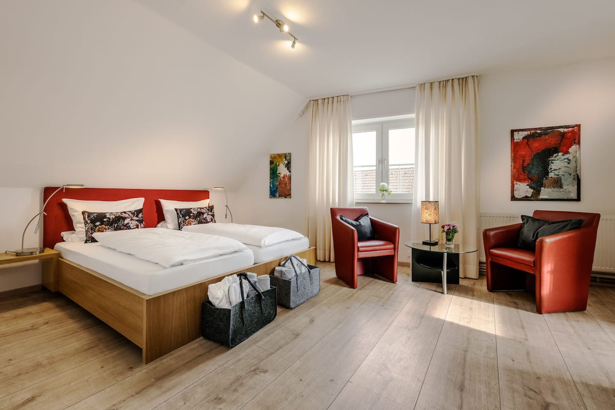 A modern hotel room with a wooden bed, two red chairs, a small table with a lamp, and a window with curtains.
