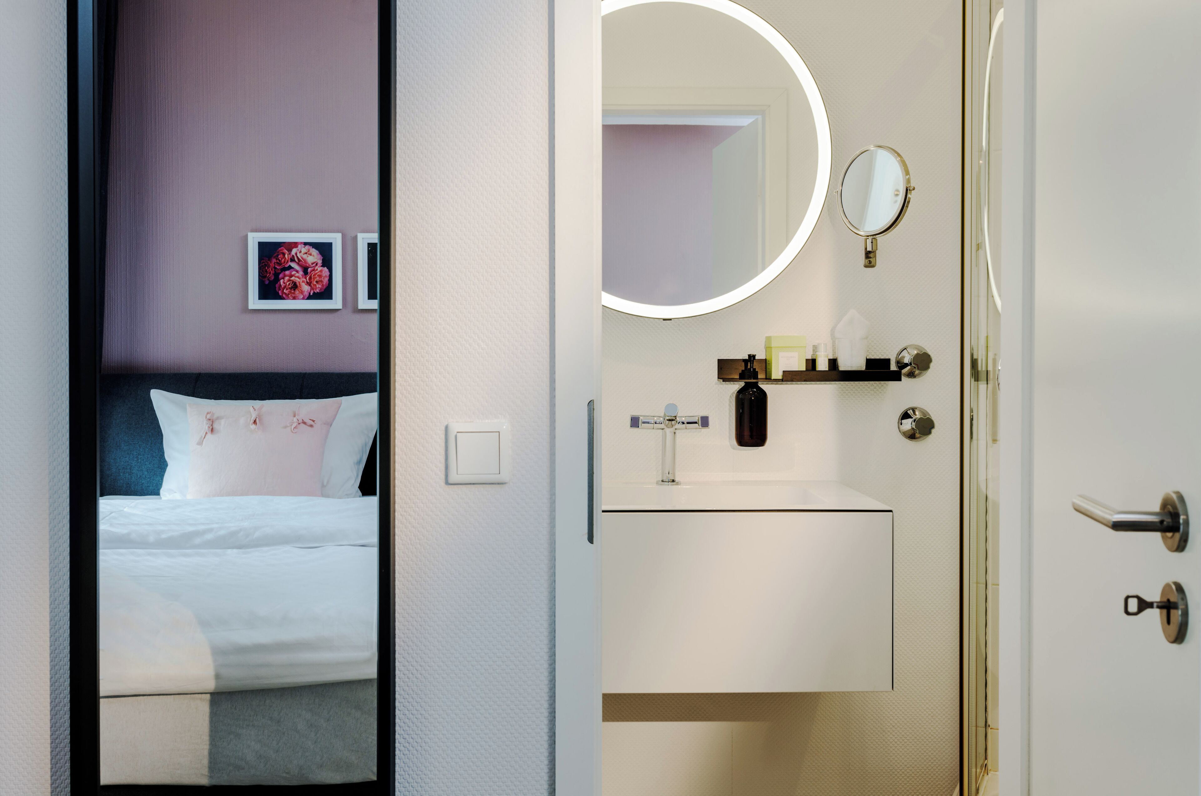 A hotel room with a bed, a small bedside table, a mirror, and a wall-mounted shelf with toiletries.