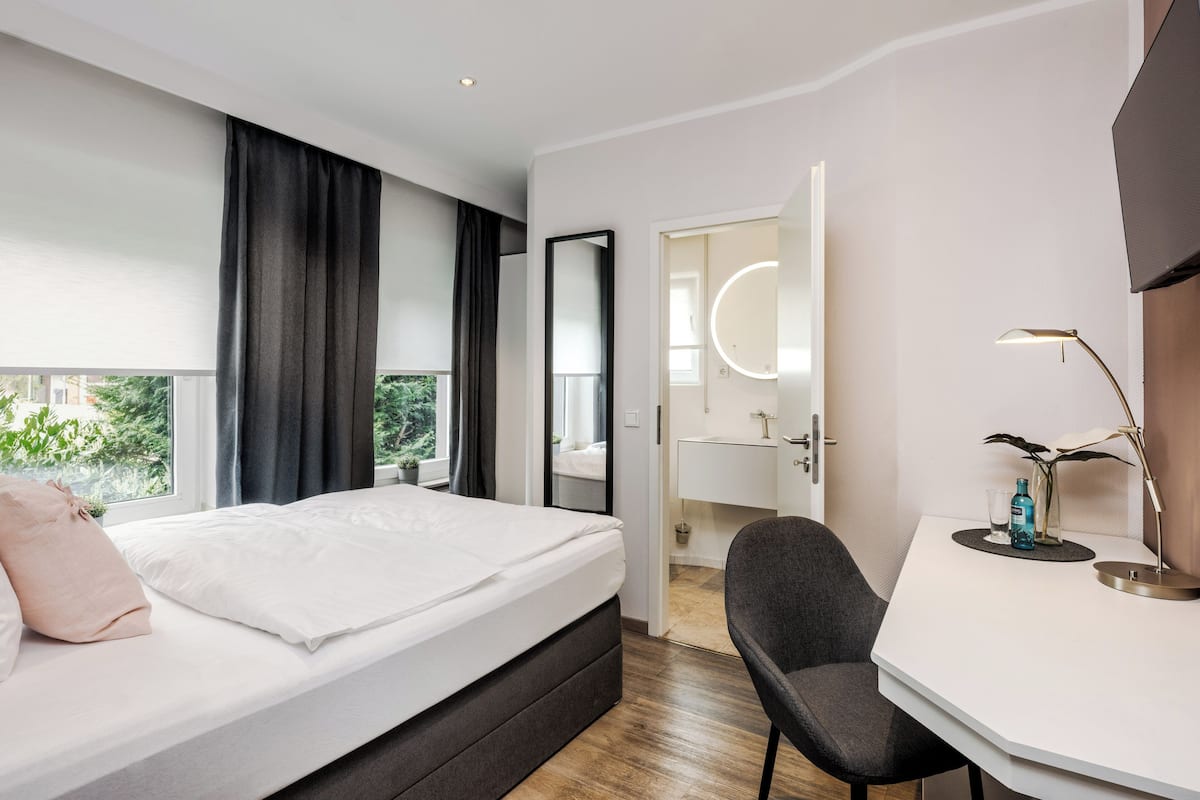 A modern hotel room with a large bed, a desk, a chair, a mirror, and a view of greenery outside.