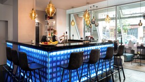 Un área de bar moderna con una barra iluminada con neón, múltiples lámparas colgantes y vista a la calle.