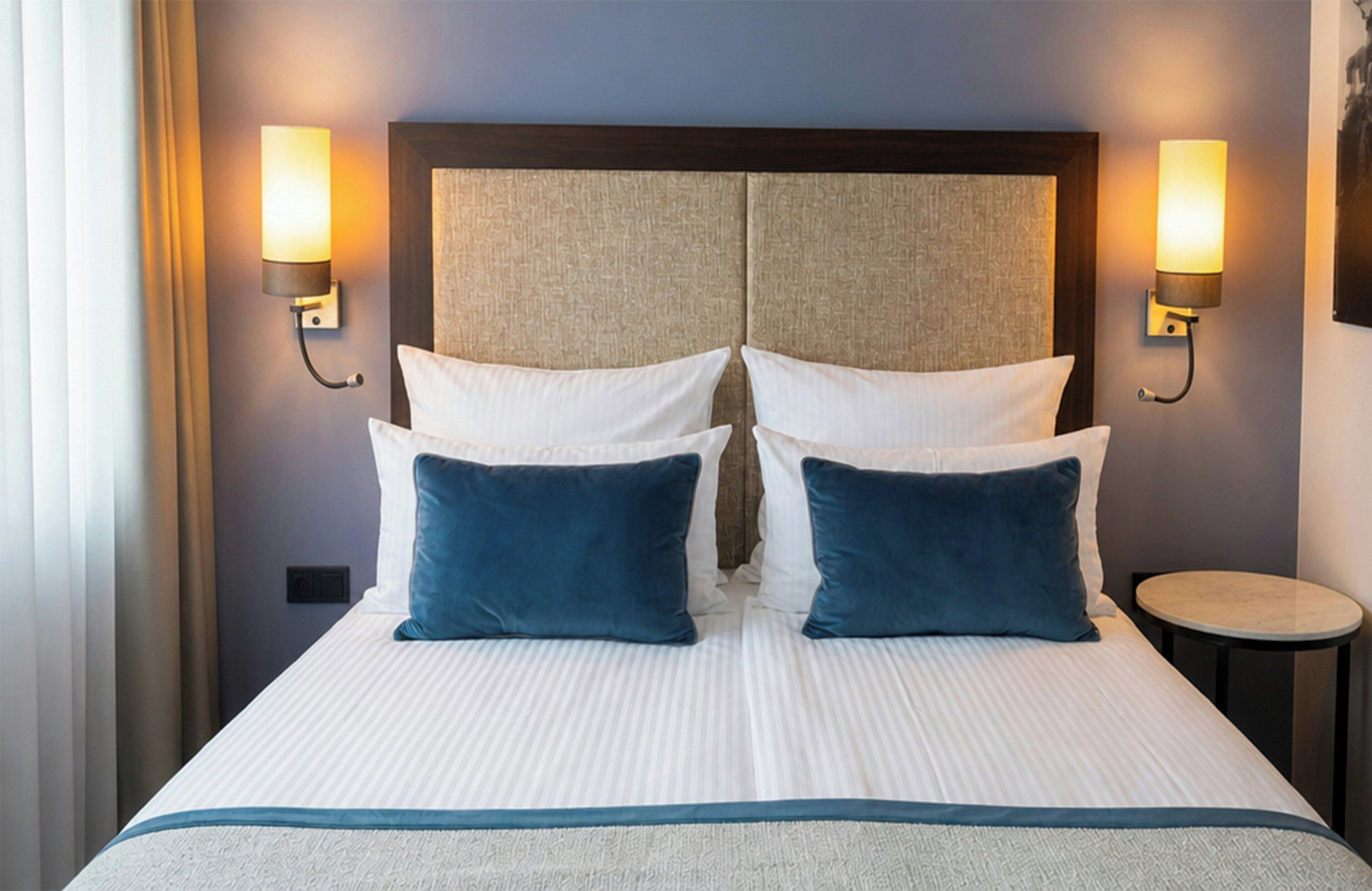 Ein Hotelzimmer mit einem großen Bett, zwei Wandlampen, einem runden Beistelltisch und einem Kopfteil mit Struktur.