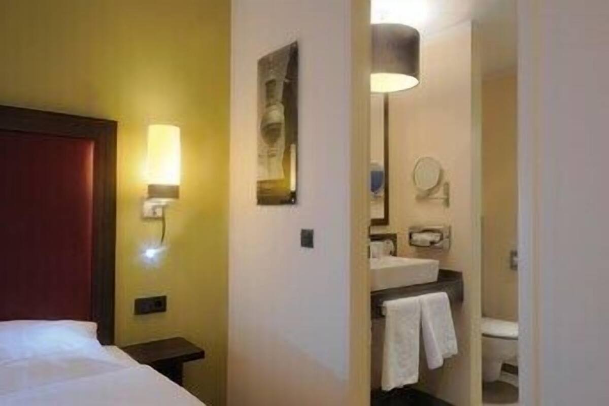 A hotel room with a bed, a nightstand, a wall-mounted lamp, a mirror, a sink, and a towel rack.
