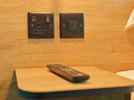A remote control on a wooden table with a wall-mounted electrical outlet and switch.