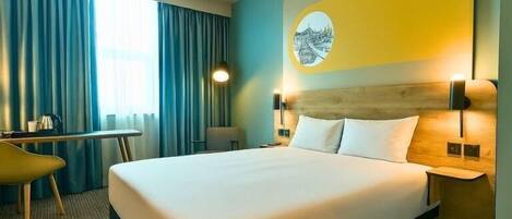 A hotel room with a large bed, a desk, and a chair. There is a window with curtains, a wall-mounted lamp, and a round picture on the wall.