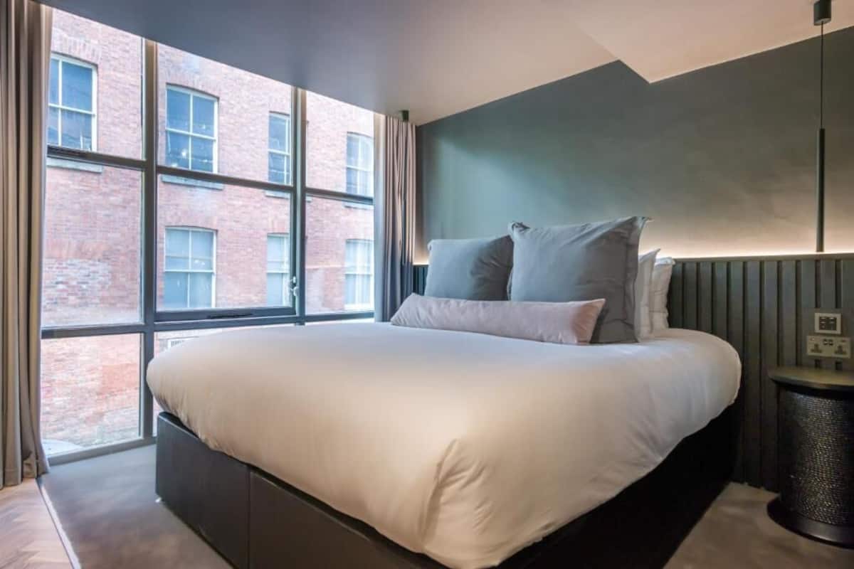 A modern bedroom with a large bed, a view of a brick building through a window, and a dark wall.