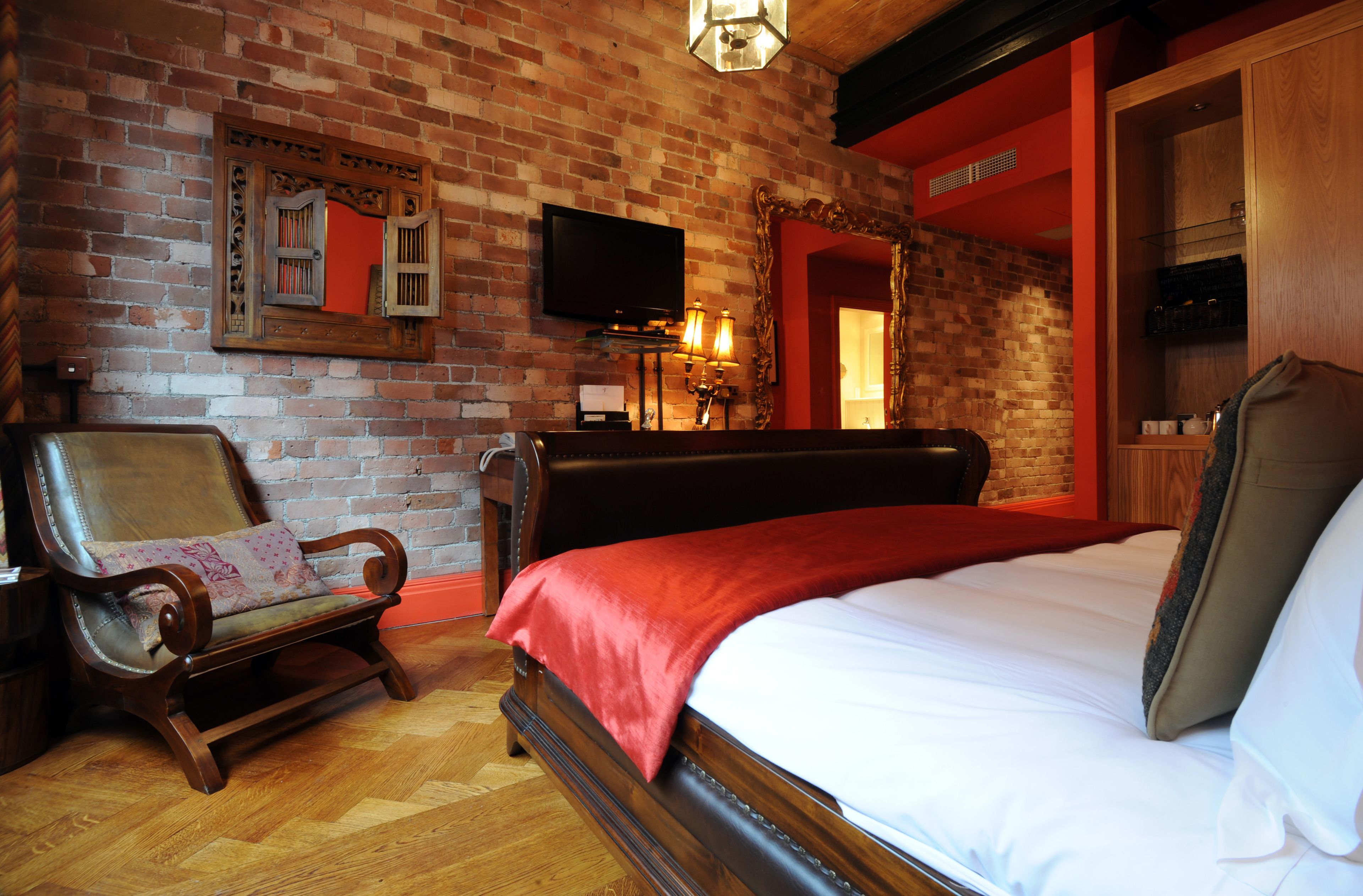 A room with a brick wall, a wooden bed, a bench, a television, and a mirror.