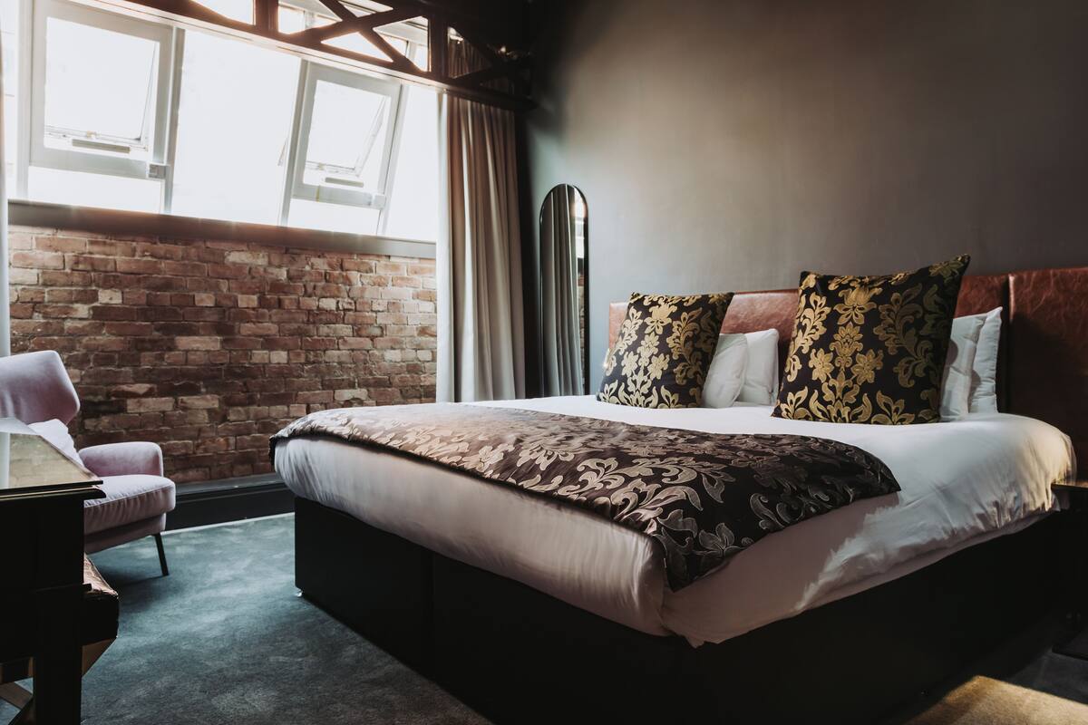A hotel room with a large bed, a brick wall, a window, and a modern chair.