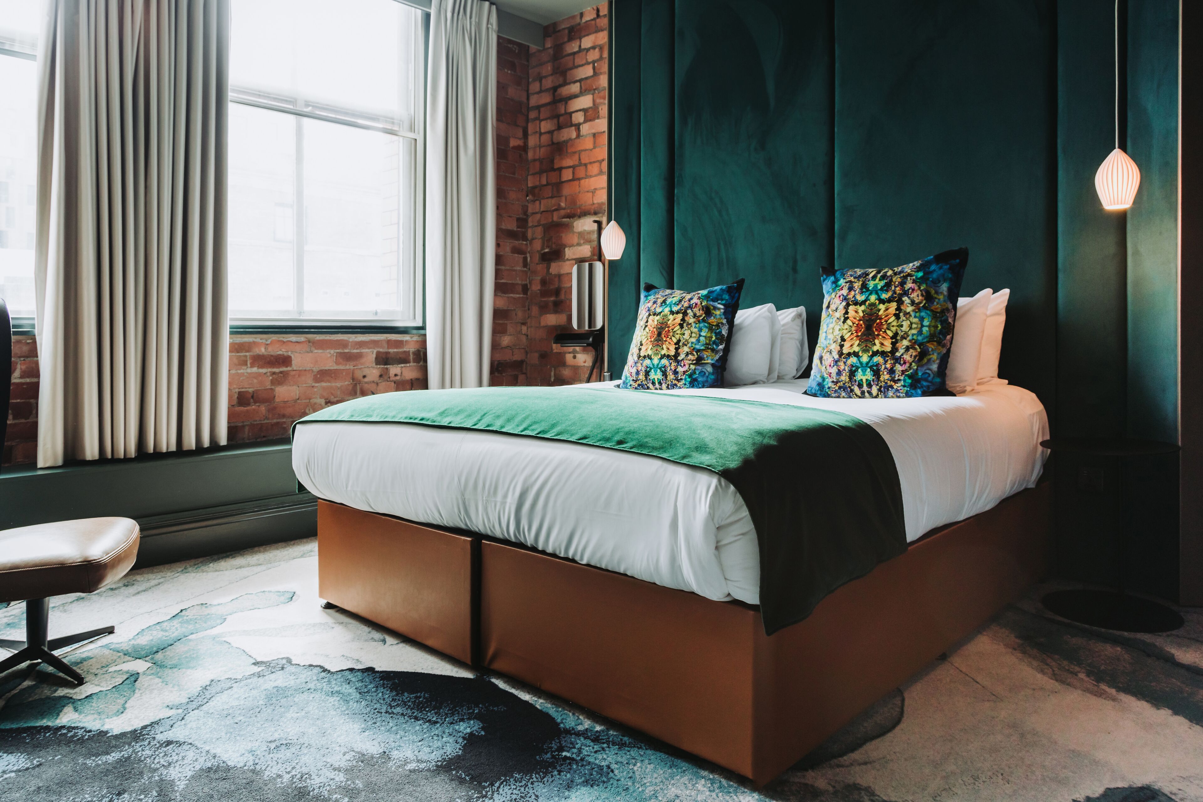 A modern bedroom with a brick wall, a large window with curtains, a green upholstered bed, and a round table with a chair.