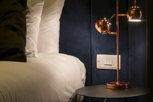 Una cama bien tendida con sábanas blancas, una mesa redonda con una lámpara de pie de cobre y un interruptor de pared.