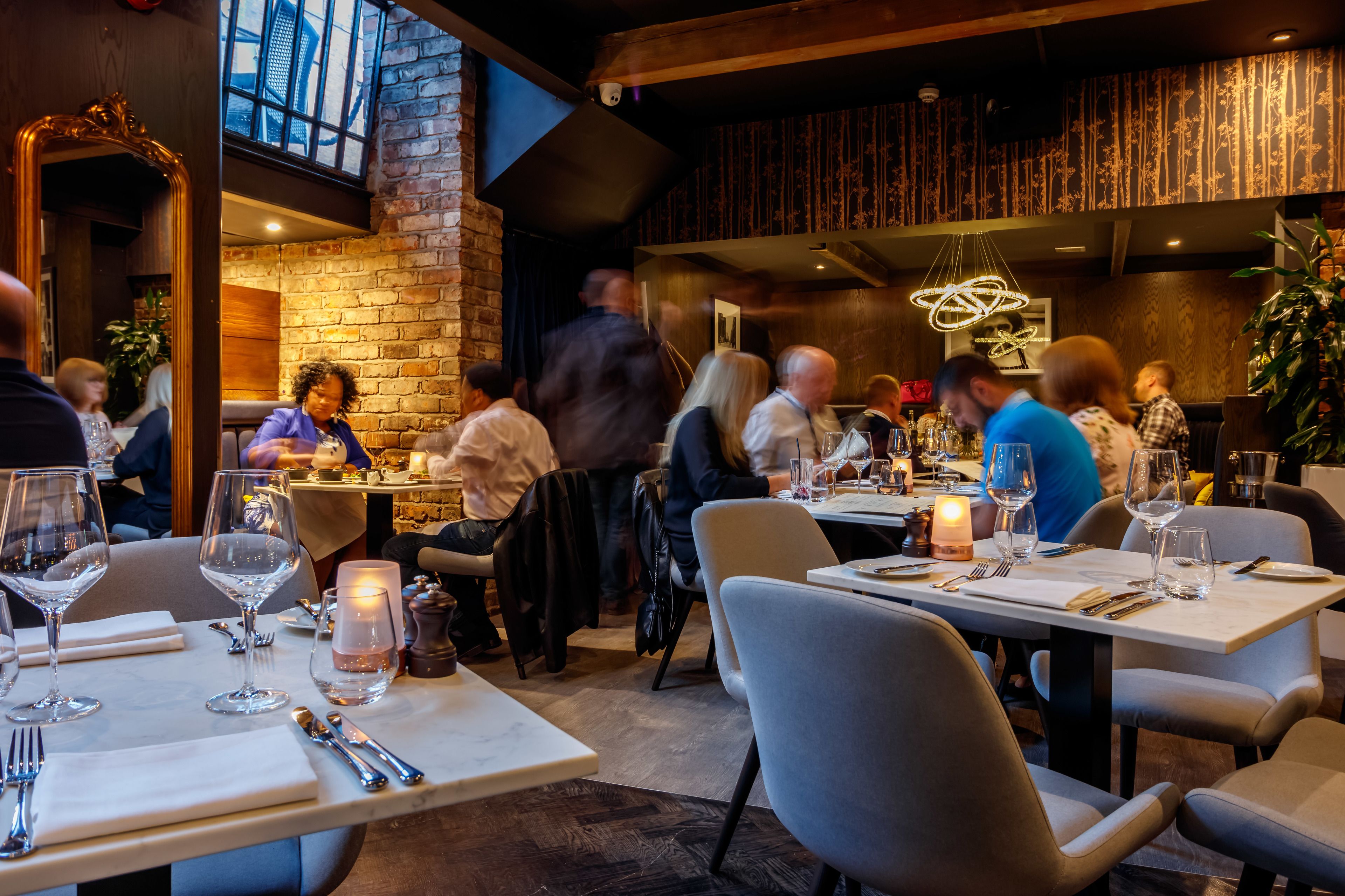 A restaurant with tables set for dining, a brick wall, and a large mirror.