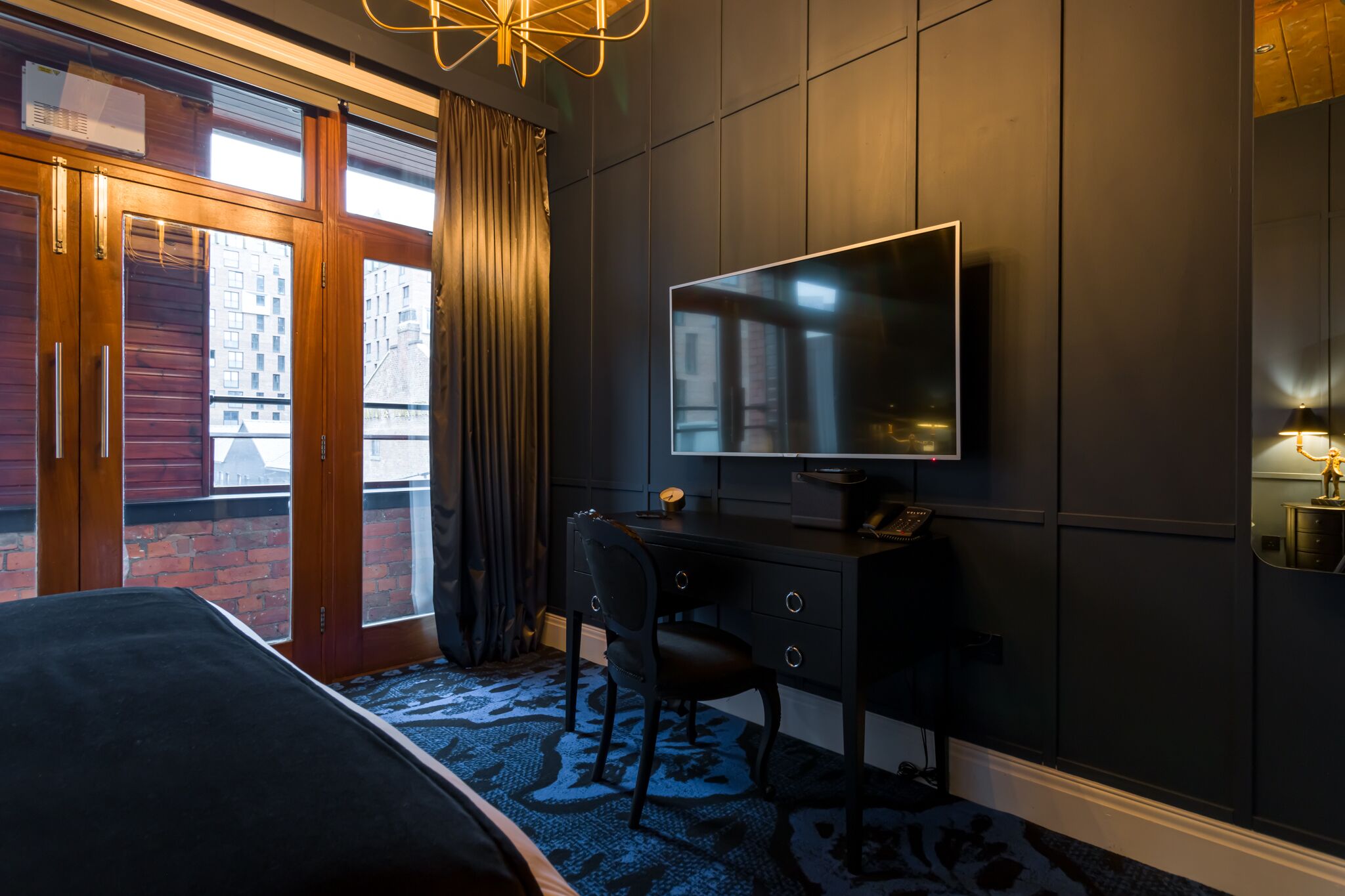 A hotel room with a large bed, a desk with a chair, a flat-screen TV, and a view of the city through the window.