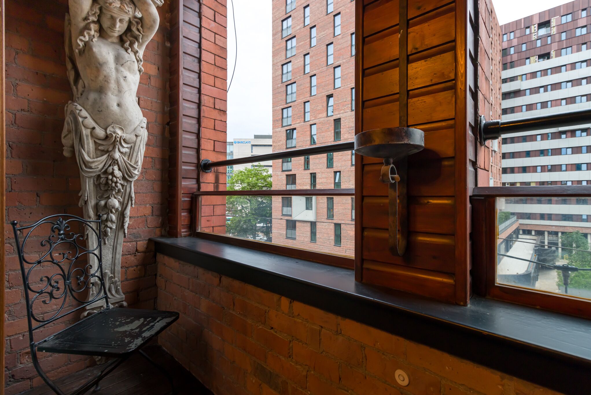 Un balcon avec un banc en fer forgé, une statue en pierre et une vue sur un bâtiment en briques.
