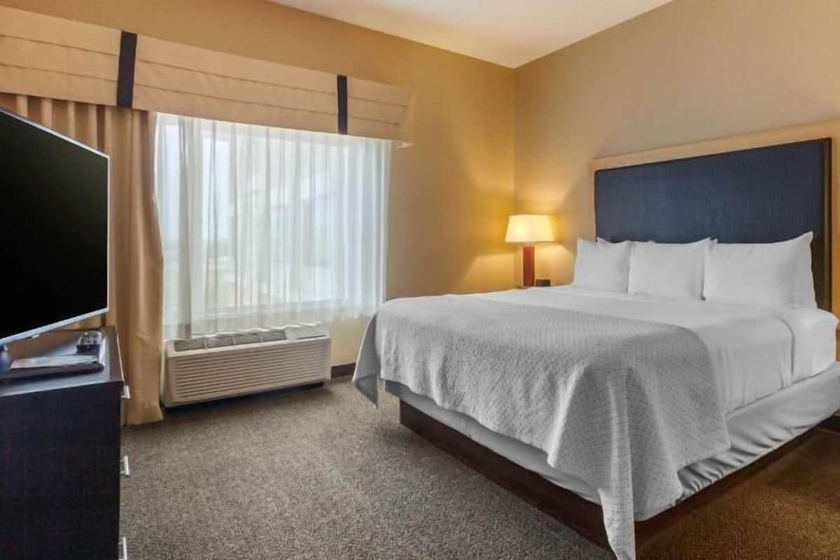 Habitación de hotel con cama, televisor, escritorio, lámpara y ventana con cortinas.