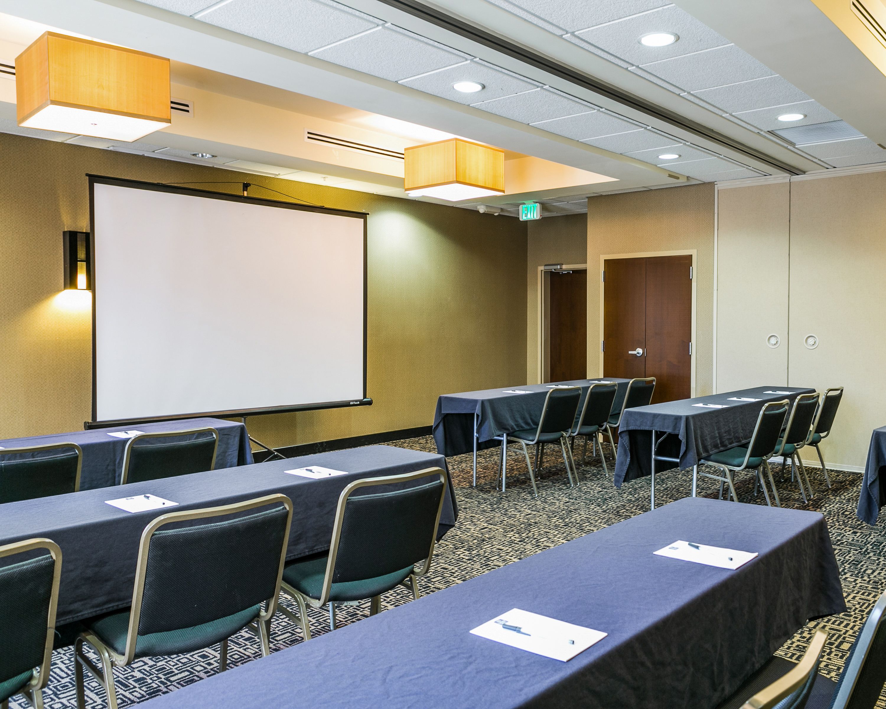 A conference room with a projector screen, tables, chairs, and a carpeted floor.