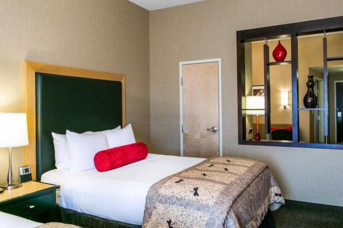 Habitación de hotel con cama, mesita de noche, lámpara y espejo con un jarrón rojo.