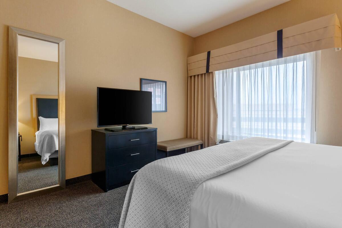 Una habitación de hotel con cama, televisor en una cómoda, ventana con cortinas y un espejo.