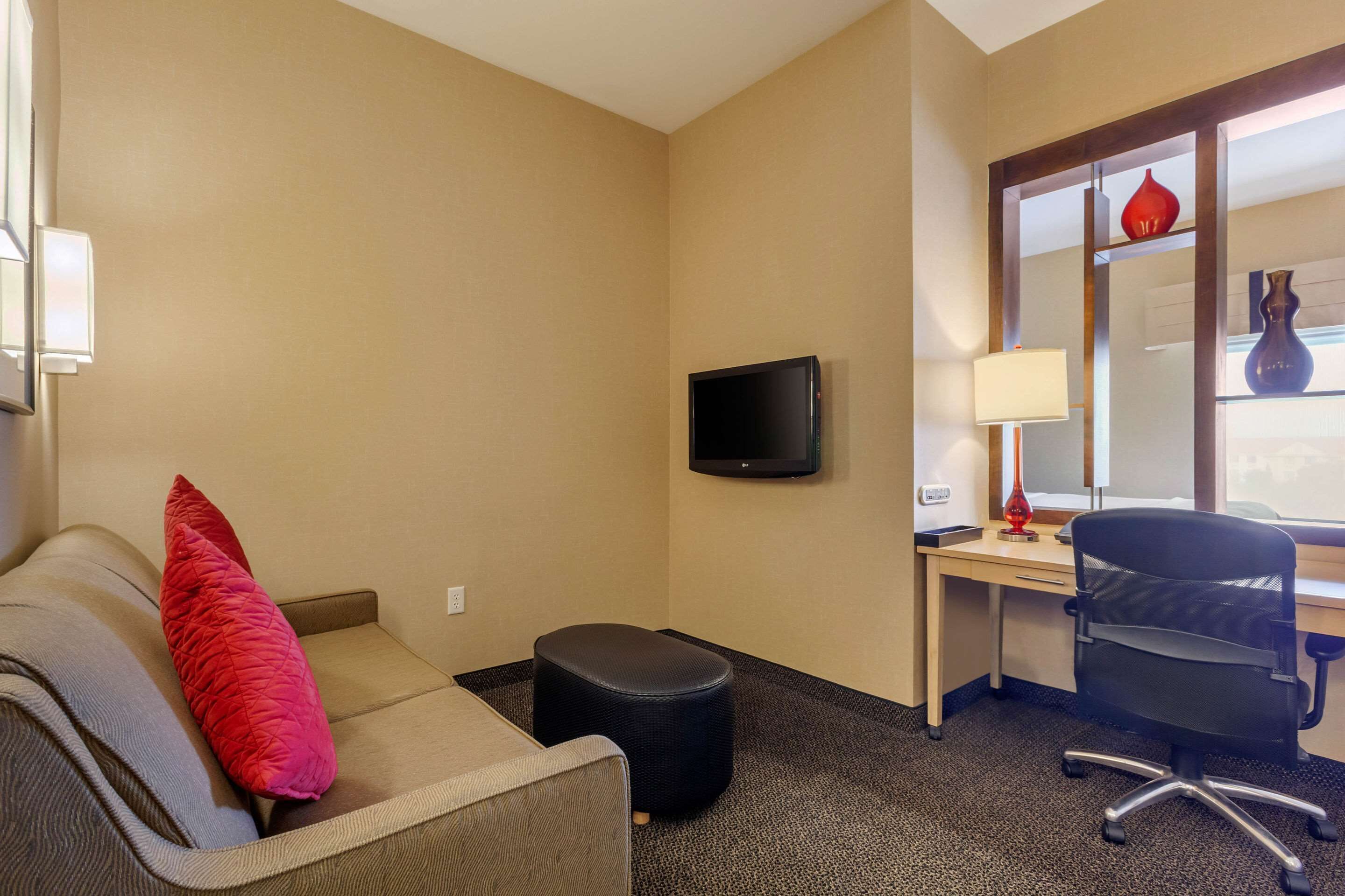 A hotel room with a sofa, ottoman, desk, chair, TV, lamp, and a window with a view.