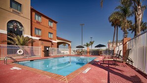 Une piscine dotée d’une terrasse carrelée de carreaux rouges, entourée d’une clôture blanche et jouxtant un bâtiment à la façade en briques et aux grandes fenêtres.