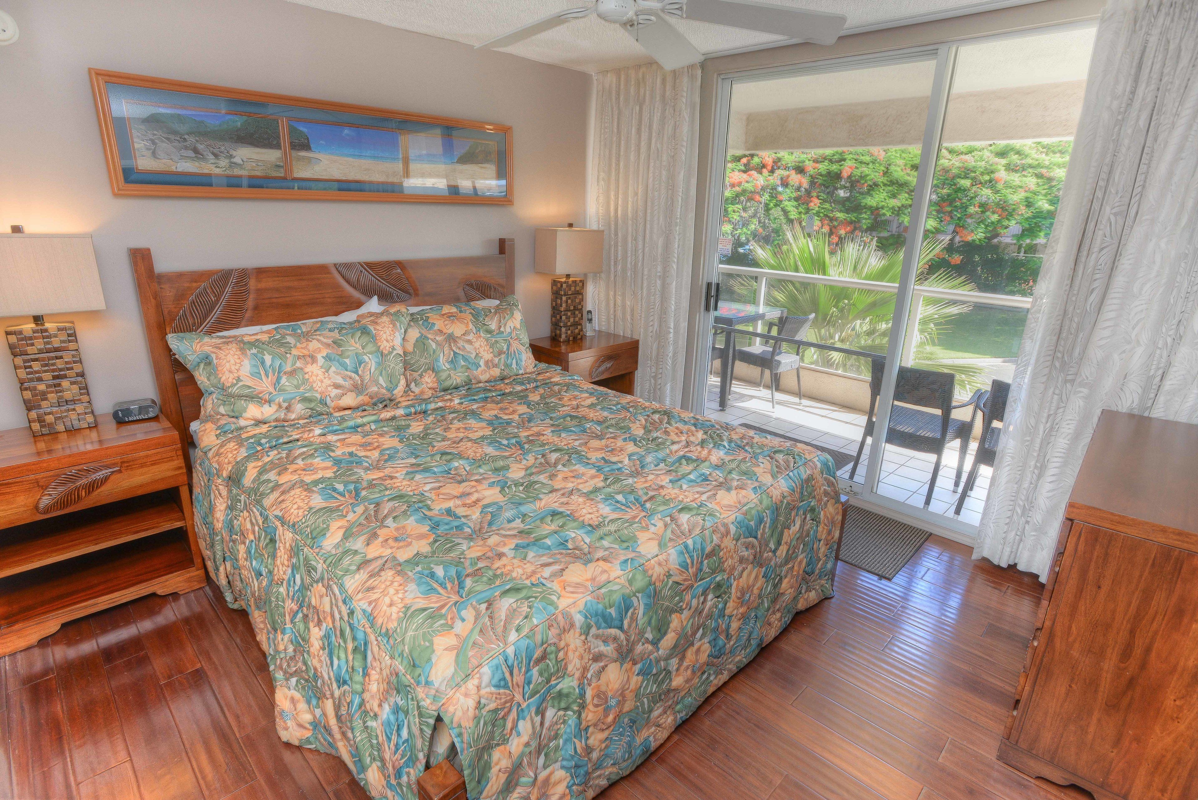Une chambre à coucher avec un grand lit, une tête de lit en bois, une table de chevet, une lampe, un tableau encadré au mur et une vue sur un balcon aménagé avec du mobilier d’extérieur.