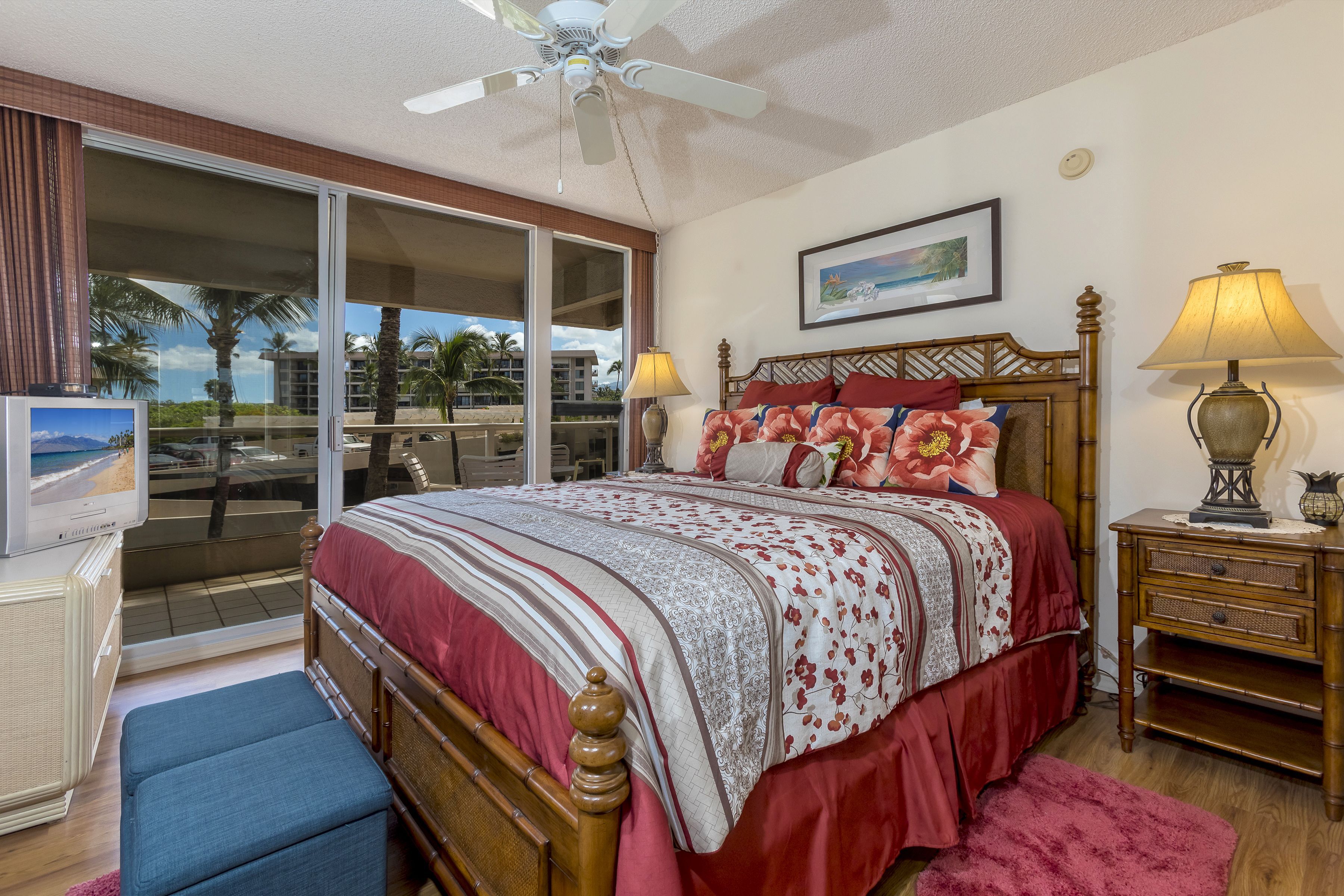 A bedroom with a bed, a television, a ceiling fan, a nightstand, and a view of the outdoors.