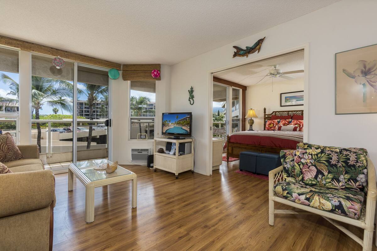 Un salón amplio con un gran ventanal corredera que da a un balcón, una televisión, una cama con cabecero rojo y una silla de mimbre.