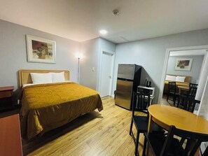 A hotel room with a bed, a refrigerator, a dining table with chairs, and a ceiling light.