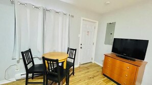 Una habitación con piso de madera, una mesa redonda con cuatro sillas, un televisor sobre un mueble de madera y cortinas blancas.